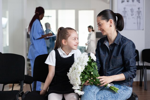 Menina sorridente conversando com a mãe na sala de espera da clínica médica. Membros da família felizes esperando para ver o parente hospitalizado no sanatório. Sala de visita do hospital ocupado.