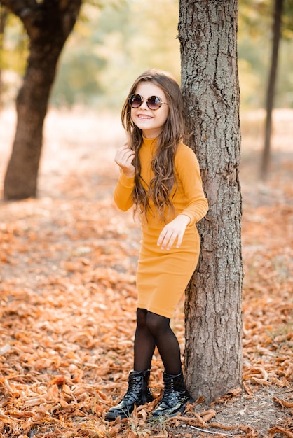 Menina sorridente de 4 a 5 anos usa vestido de malha amarela e óculos de sol posando no parque ao ar livre