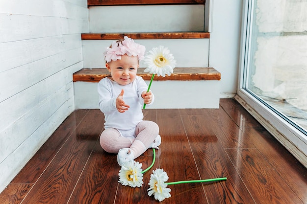 Menina sorridente de um ano de idade usando coroa de primavera no chão na sala de estar de luz brilhante perto da janela e brincando com flores gerbera Criança feliz brincando em casa Conceito de infância