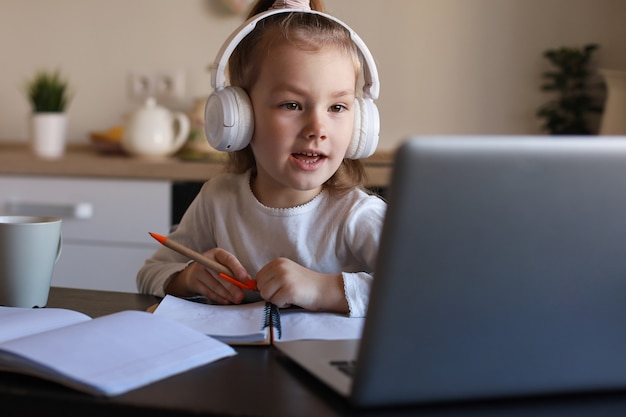 Menina sorridente em fones de ouvido manuscrita estudo on-line usando o laptop em casa, criança pequena feliz fofa em fones de ouvido fazer aula de web sobre Internet ou aula no PC.