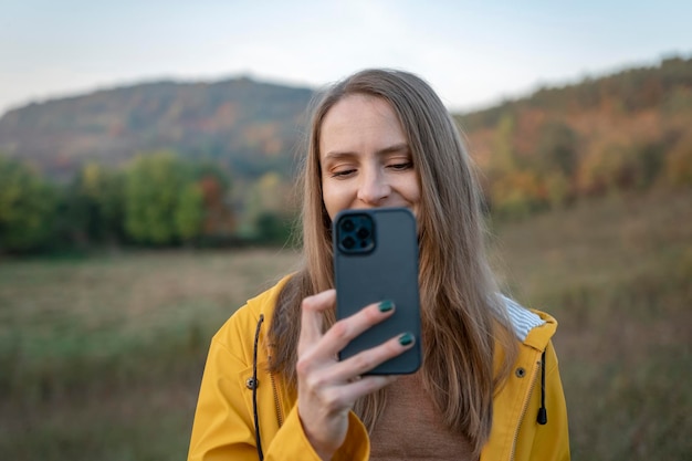 Menina sorridente em jaqueta amarela usa smartphone enquanto caminhava no dia de outono na natureza Jovem fotografa bela paisagem com telefone celular