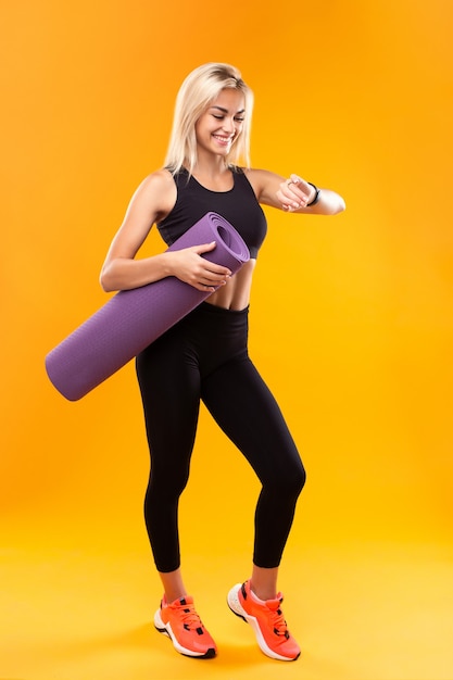 Menina sorridente em roupas esportivas com um relógio inteligente e um tapete nas mãos em um fundo amarelo