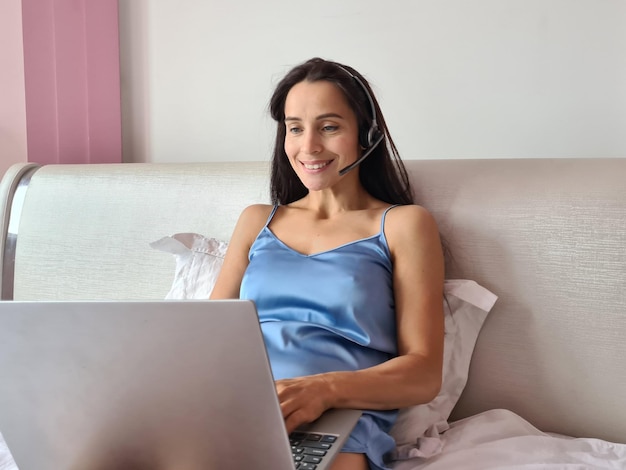 Menina sorridente em um fone de ouvido sem fio está na cama estudando online no laptop