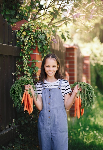 Menina sorridente em um jardim segura dois cachos de cenouras frescas vertical