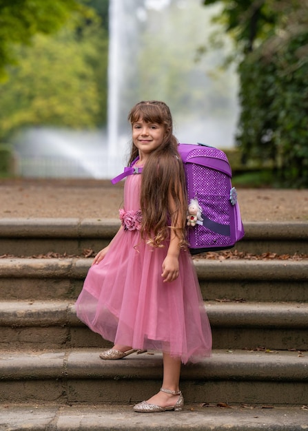 Foto menina sorridente em um lindo vestido rosa com cabelos longos e uma mochila vai para a escola