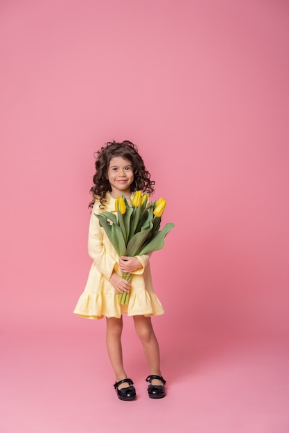 Menina sorridente em um vestido amarelo sobre fundo rosa studio. Criança feliz alegre com buquê de flores de tulipas.