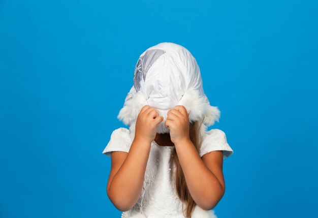 Menina sorridente em um vestido branco e um chapéu de pele branca de inverno se diverte em um fundo azul.