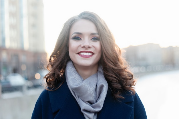 Menina sorridente entusiasmada com cachos brilhantes, posando na cidade à luz do sol. Retrato ao ar livre do close-up da senhora encantadora na moda outono trincheira com sorriso maravilhoso e grandes olhos azuis.