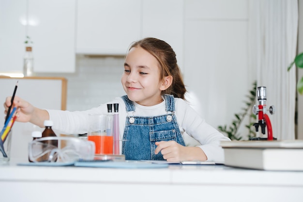 Menina sorridente fazendo projeto de ciências em casa, pegando giz de cera. ela tem vidros de química com líquidos coloridos e microscópio