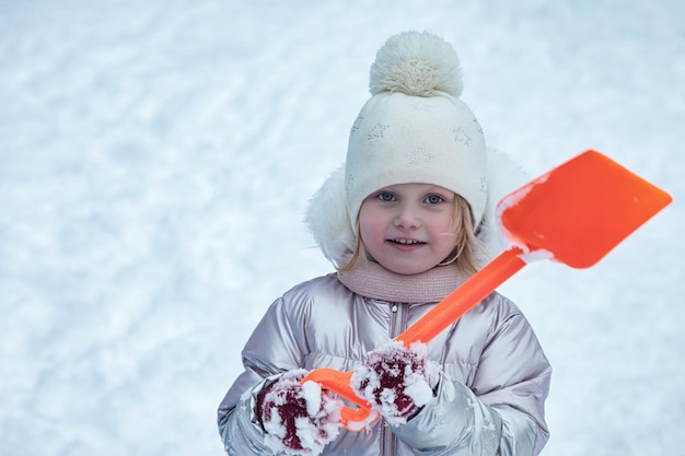 Menina sorridente feliz com pá de crianças no playground de neve de inverno congelado em roupas quentes Criança bonita se divertindo
