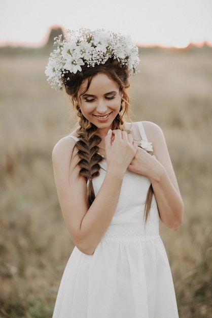 Menina sorridente feliz com tranças e guirlanda floral em vestido branco no estilo boho no verão ao ar livre