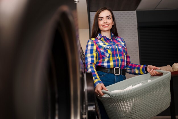 Menina sorridente fica na lavanderia e segura uma cesta de roupa