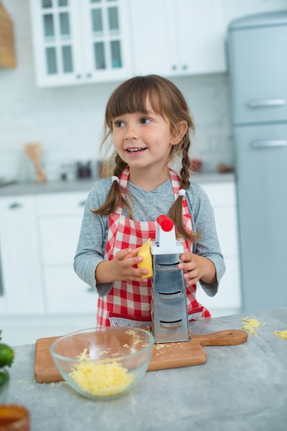 Menina sorridente fofa com rabo de cavalo e avental de cozinha quadriculado esfrega queijo em um ralador, ajuda a cozinhar pizza na cozinha.