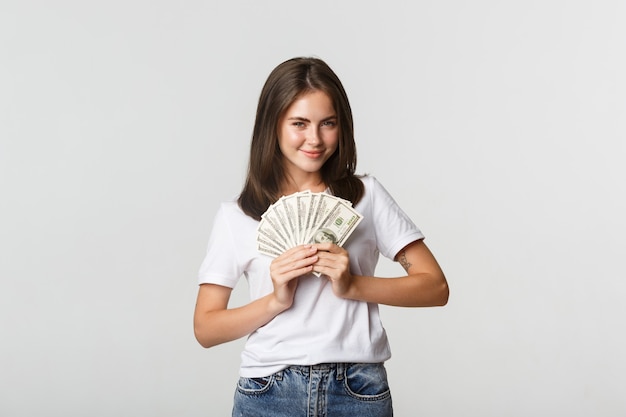 Menina sorridente gananciosa olhando astuta e segurando dinheiro, de pé branco.