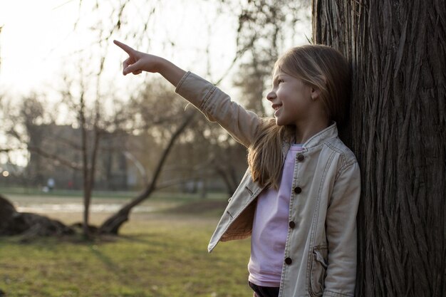 Menina sorridente no parque, encostada em uma árvore, mostrando algo com o dedo estendido.