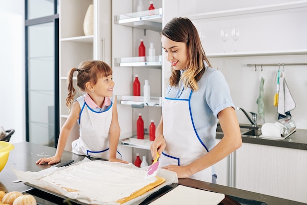 Menina sorridente, olhando para a mãe cobrindo o bolo com uma deliciosa cobertura de creme