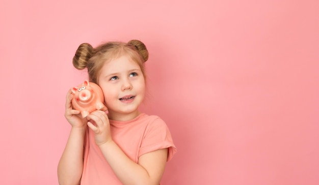 Menina sorridente segurando o cofrinho no fundo rosa Planejando finanças e economizando o conceito de dinheiro