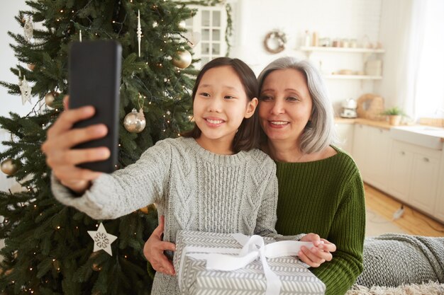 Menina sorridente segurando seu telefone celular e tirando uma foto junto com a mãe perto da árvore de natal