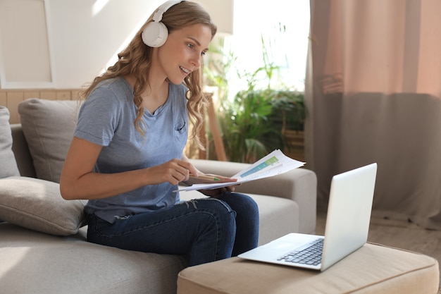 Foto menina sorridente, senta-se perto do sofá, assistindo ao webinar no laptop. estudo de mulher jovem feliz em curso distante online.