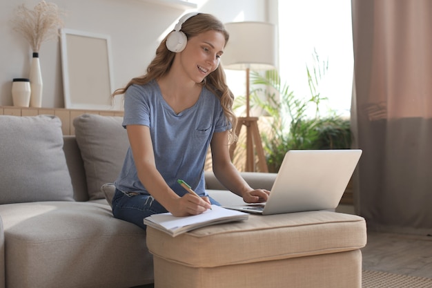 Menina sorridente, senta-se perto do sofá, assistindo ao webinar no laptop. Estudo de mulher jovem feliz em curso distante online.