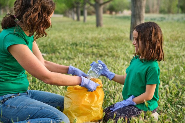 Menina sorridente sentada na grama verde com sua mãe coletando plásticos no dia da terra