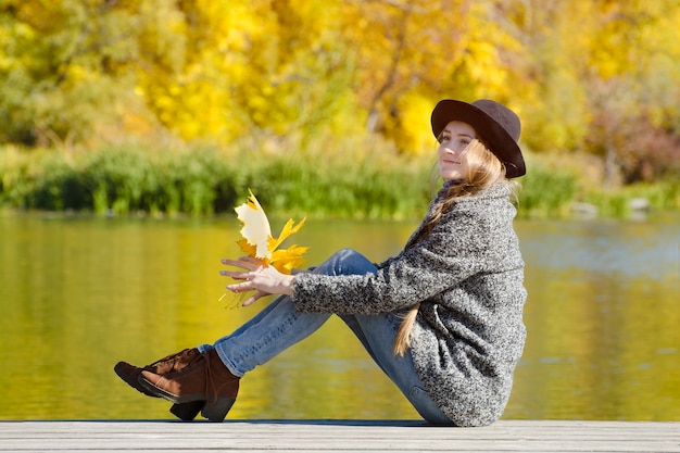 Menina sorridente sentada no cais com folhas de outono nas mãos. Dia ensolarado