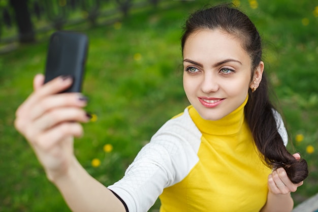 Menina sorridente tirando foto com a câmera do smartphone ao ar livre. selfie