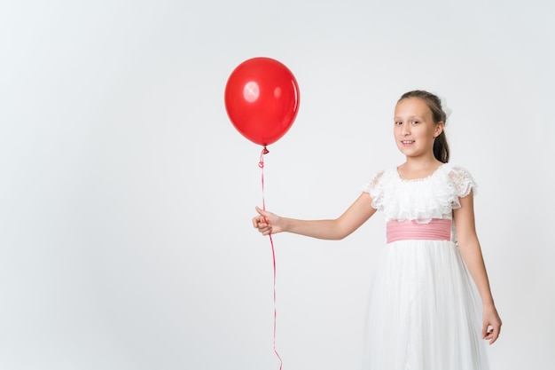 Menina sorridente vestida com um longo vestido branco segurando um balão vermelho na mão sorrindo olhando para a câmera