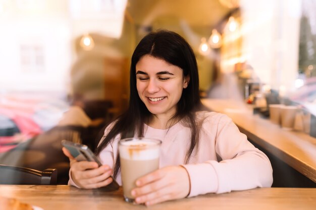 Menina sorrindo, bebe café no café e lendo o telefone.