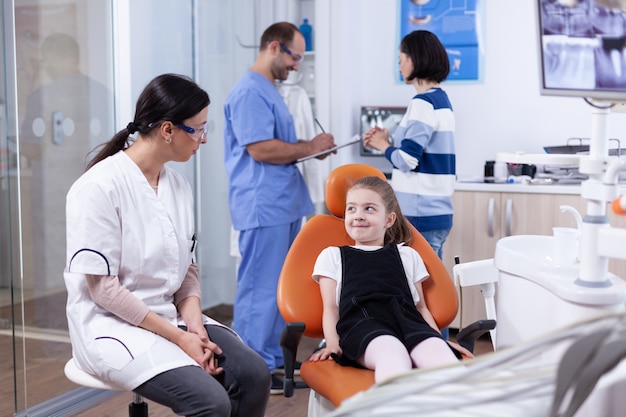 Menina sorrindo para o dentista sentado na cadeira, esperando o tratamento de dentes. Criança com a mãe durante o exame de dentes com stomatolog sentado na cadeira.