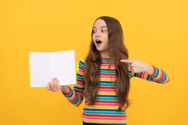 Menina surpreendida aponta o dedo para o livro aberto para o fundo amarelo do espaço de cópia, apontando.