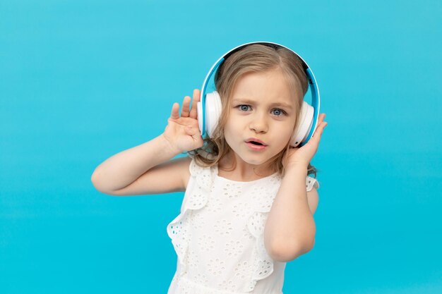 Menina surpresa com fones de ouvido ouvindo música em um vestido branco de algodão em um fundo azul no espaço do estúdio para texto