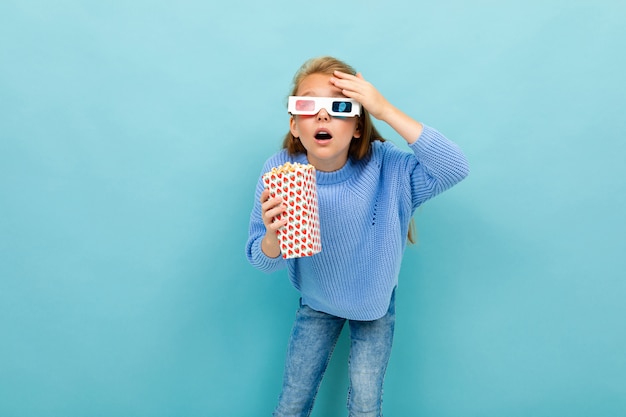 Menina surpresa em óculos de filme está segurando pipoca nas mãos na luz azul