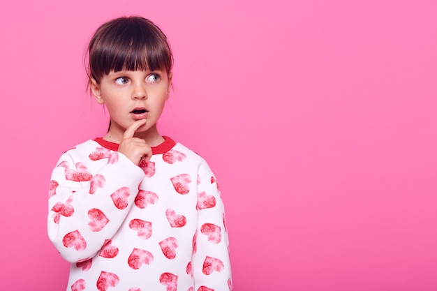 Menina surpresa olhando para o lado com a boca bem aberta e o dedo perto da boca, copie o espaço, vestindo um suéter, isolado sobre a parede rosa.