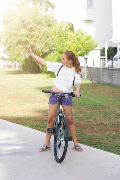 Menina tirando uma selfie com um telefone inteligente enquanto está de pé com uma bicicleta na calçada