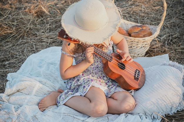 Menina tocando ukulele sentada no cobertor no campo Retrato de criança escondendo o rosto sob o chapéu panamá Piquenique ao ar livre
