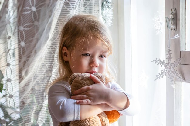 menina triste abraços e beijos ursinho de brinquedo. Criança perto da janela de luz em casa. Conceito de suporte