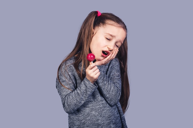 Menina triste com um pirulito vermelho nas mãos, cujos dentes doem. o conceito de desenvolvimento de cárie devido ao abuso de doces. isolado na superfície cinza