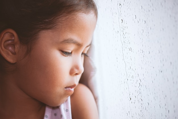 Menina triste criança asiática olhando do lado de fora pela janela no dia chuvoso