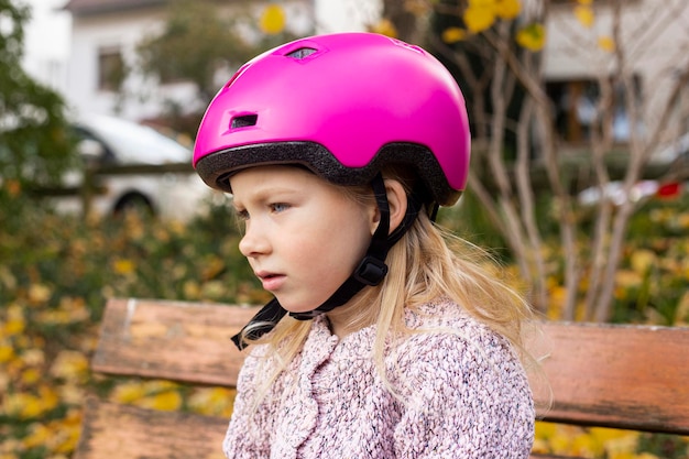 Menina triste em um capacete rosa sentado em um banco do parque