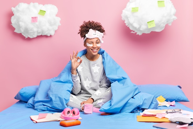 menina usa roupa de dormir sorri amplamente faz gesto de ok dá excelente gesto piscadela olhos poses pernas cruzadas na cama confortável rodeada de papéis adesivos de caderno