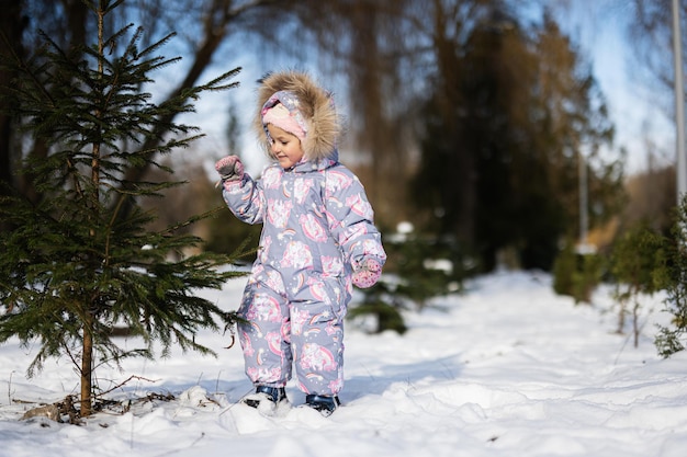 Menina usa roupa de neve infantil em um dia ensolarado e gelado de inverno perto da árvore de Natal