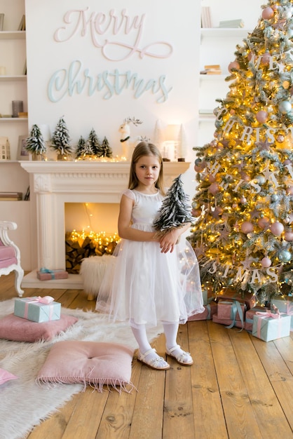 Menina, vestida com um lindo vestido branco elegante, posa perto da árvore de natal.