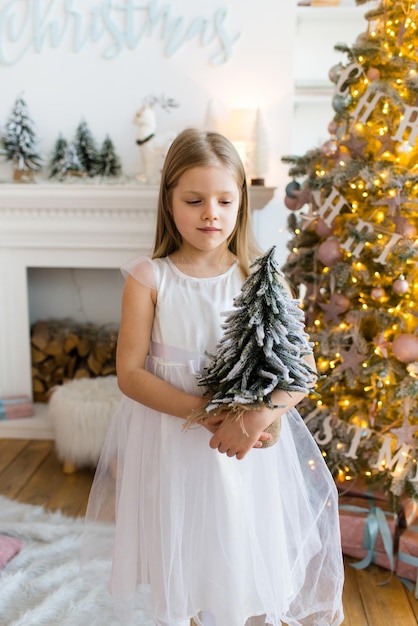 menina, vestida com um lindo vestido branco elegante, posa perto da árvore de Natal.