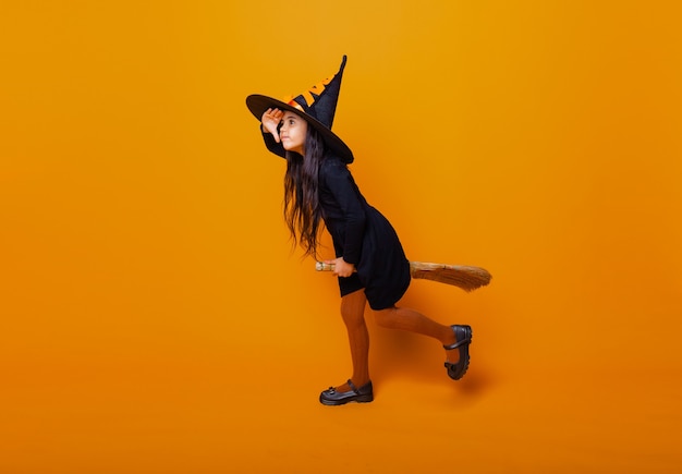 Foto menina vestida como uma bruxa de halloween em um vestido preto e um chapéu voa em uma vassoura em um fundo amarelo.