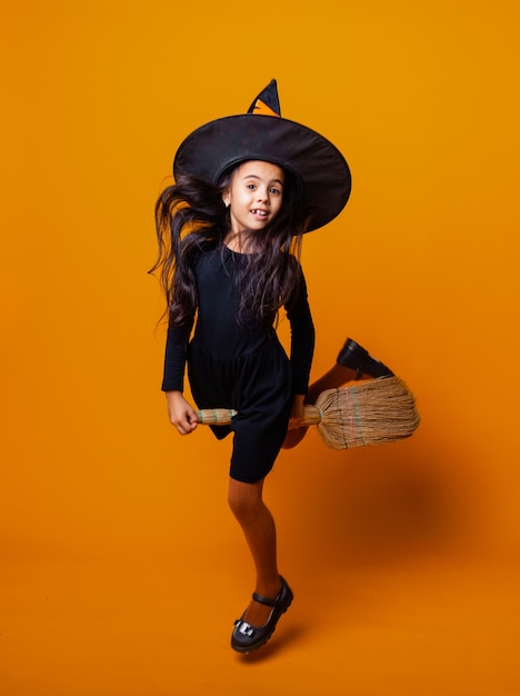 Menina vestida como uma bruxa de Halloween em um vestido preto e um chapéu voa em uma vassoura em um fundo amarelo.