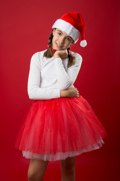 Menina vestida de Papai Noel com fundo vermelho e mão no queixo