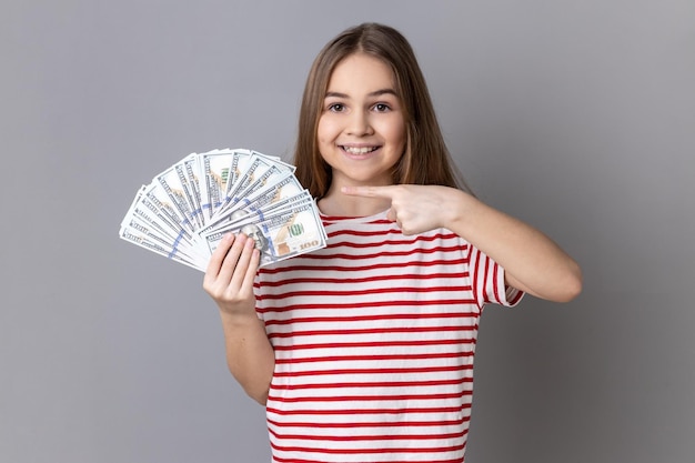 Menina vestindo apontando para fã de notas de dólar mostrando muito dinheiro e olhando para a câmera