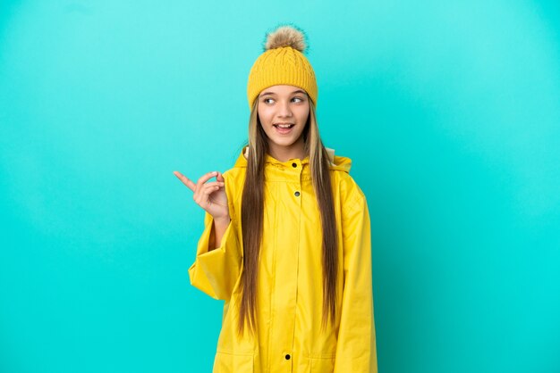 Menina vestindo um casaco à prova de chuva sobre um fundo azul isolado com a intenção de descobrir a solução enquanto levanta um dedo