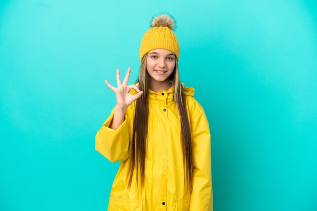 Menina vestindo um casaco à prova de chuva sobre um fundo azul isolado, mostrando um sinal de ok com os dedos